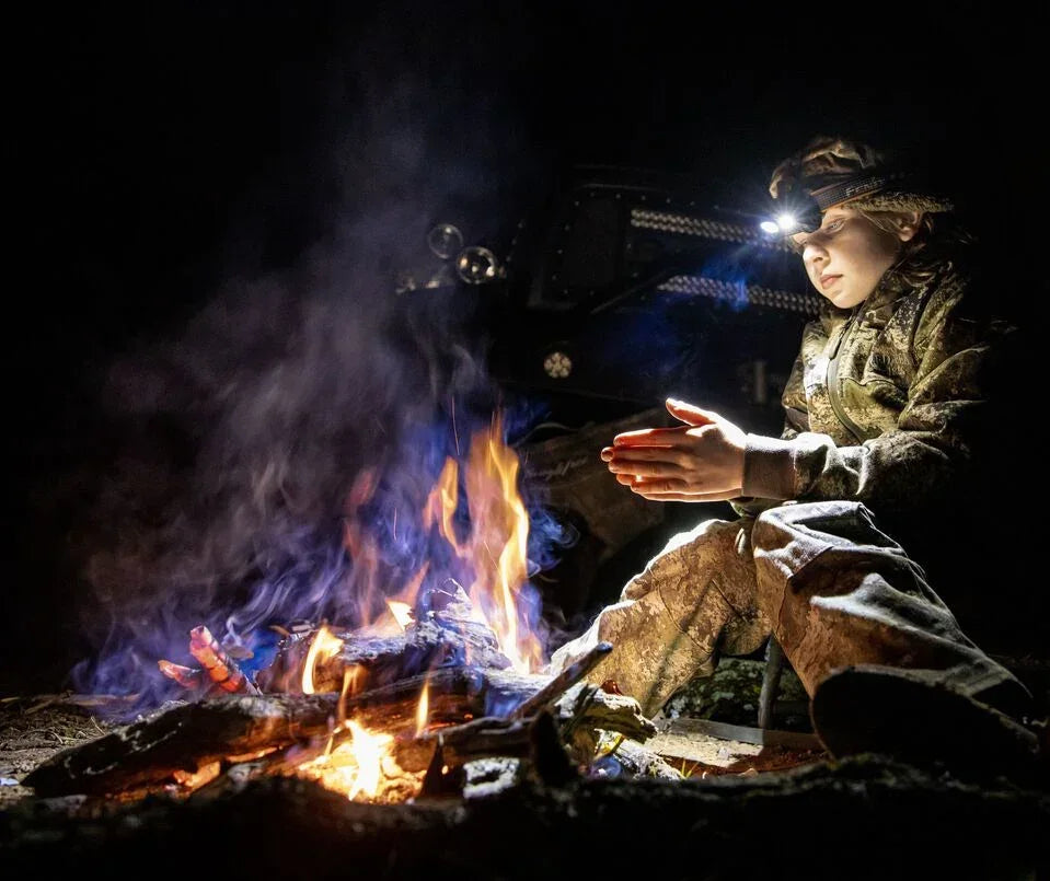 Kind in Tarnkleidung wärmt Hände am Lagerfeuer bei Nacht mit Fenix Stirnlampe in freier Natur