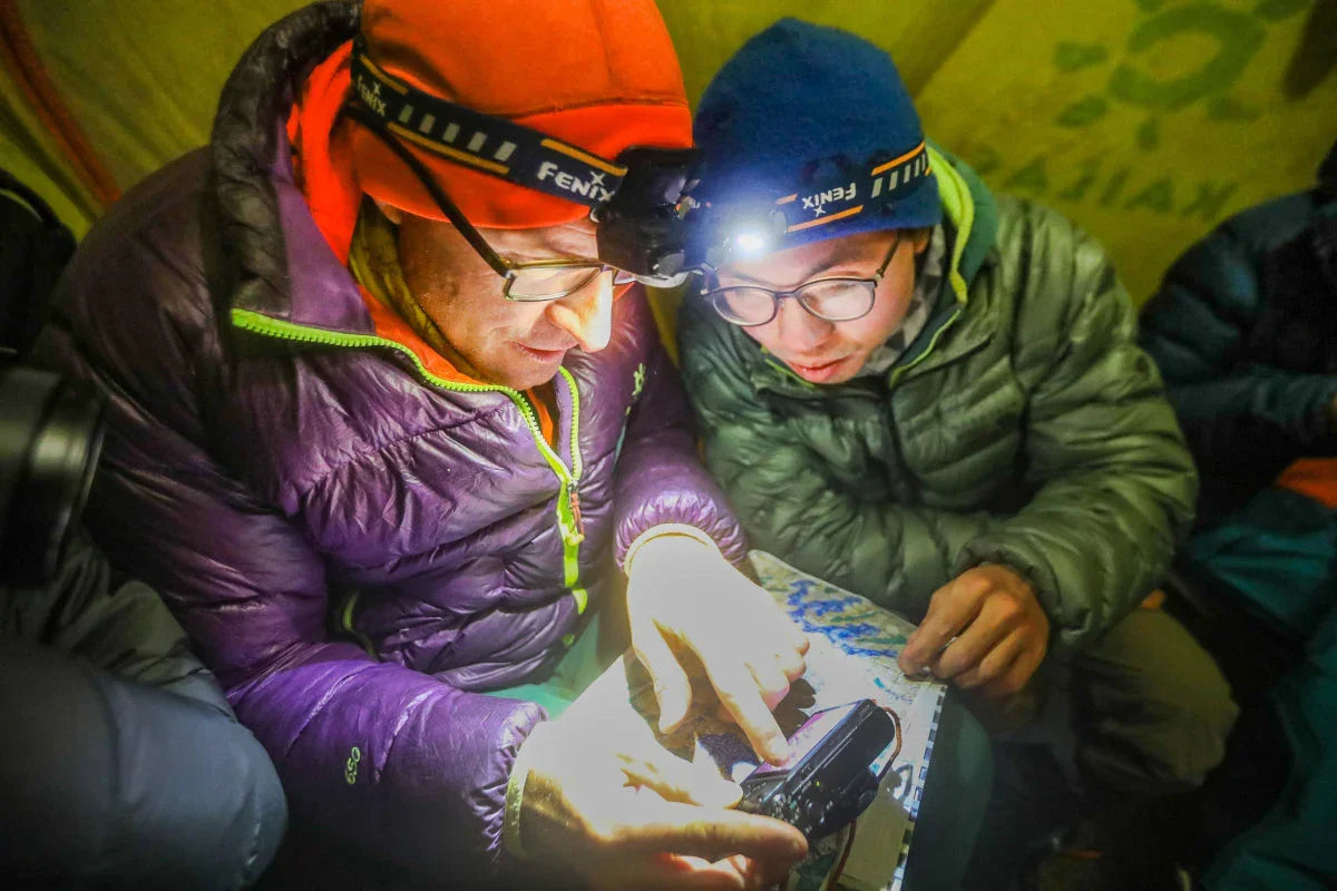 Zwei Wanderer in Outdoorjacken mit Stirnlampen lesen bei Nacht Karte und GPS-Gerät im Zelt