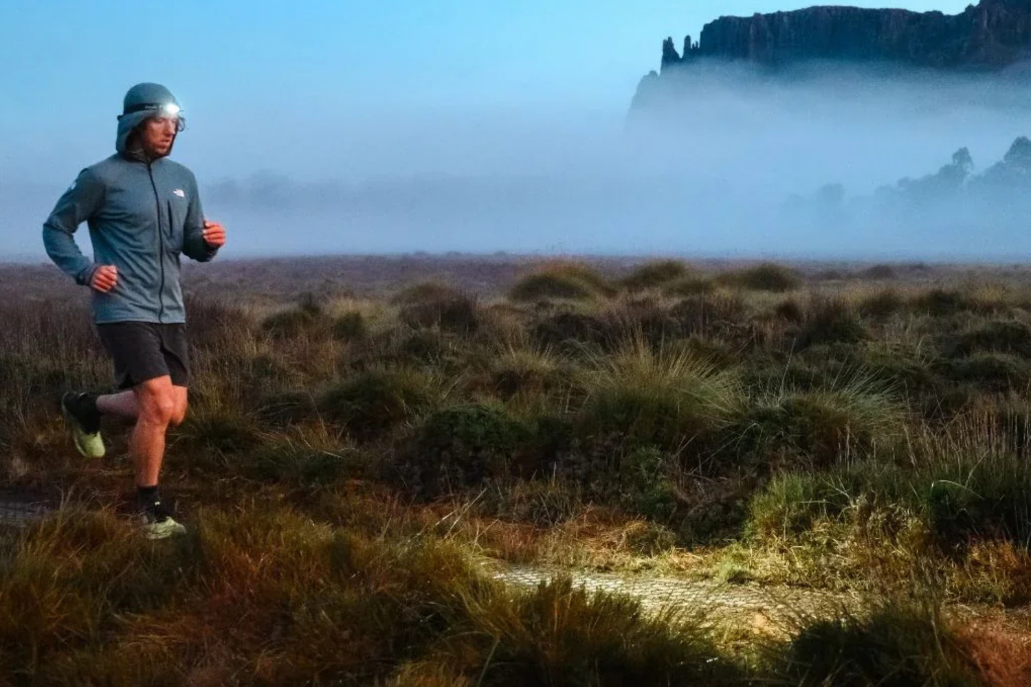 Frau läuft nachts im Gebirge mit Stirnlampe und Outdoor-Laufkleidung auf Trail mit Schneeresten