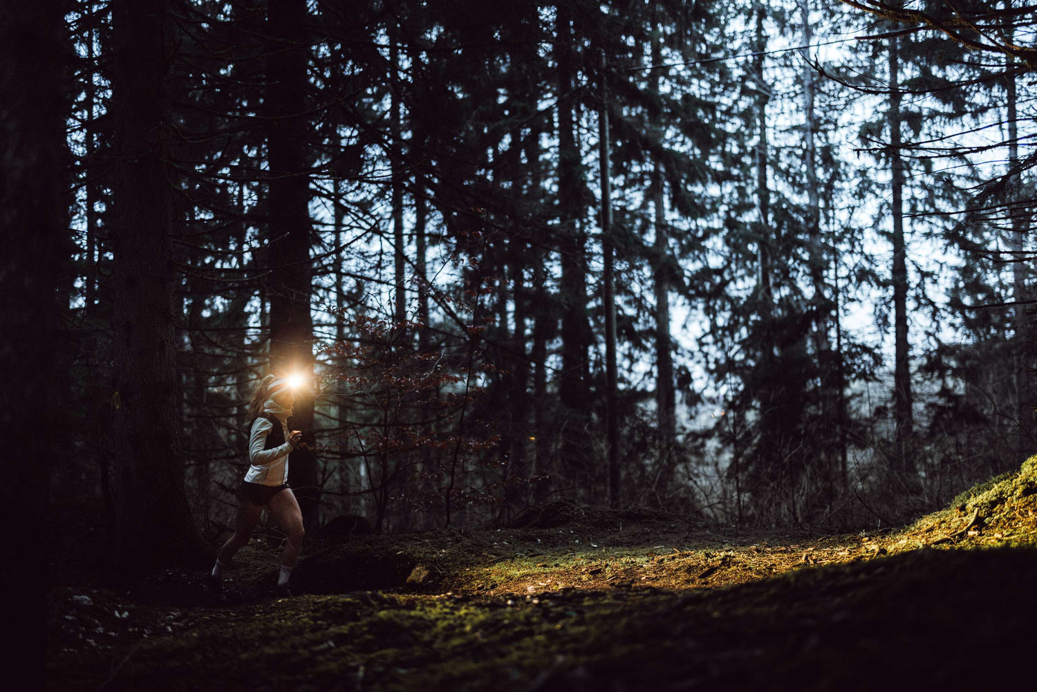 Frau läuft nachts im Gebirge mit Stirnlampe und Outdoor-Laufkleidung auf Trail mit Schneeresten