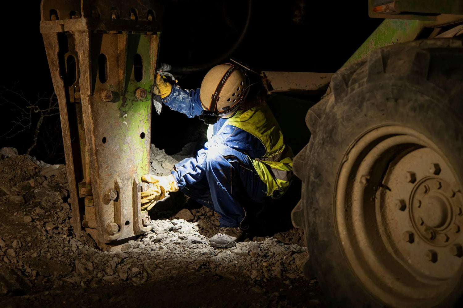Baustellenarbeiter mit Helm, Warnweste und Stirnlampe repariert Baumaschine bei Nacht