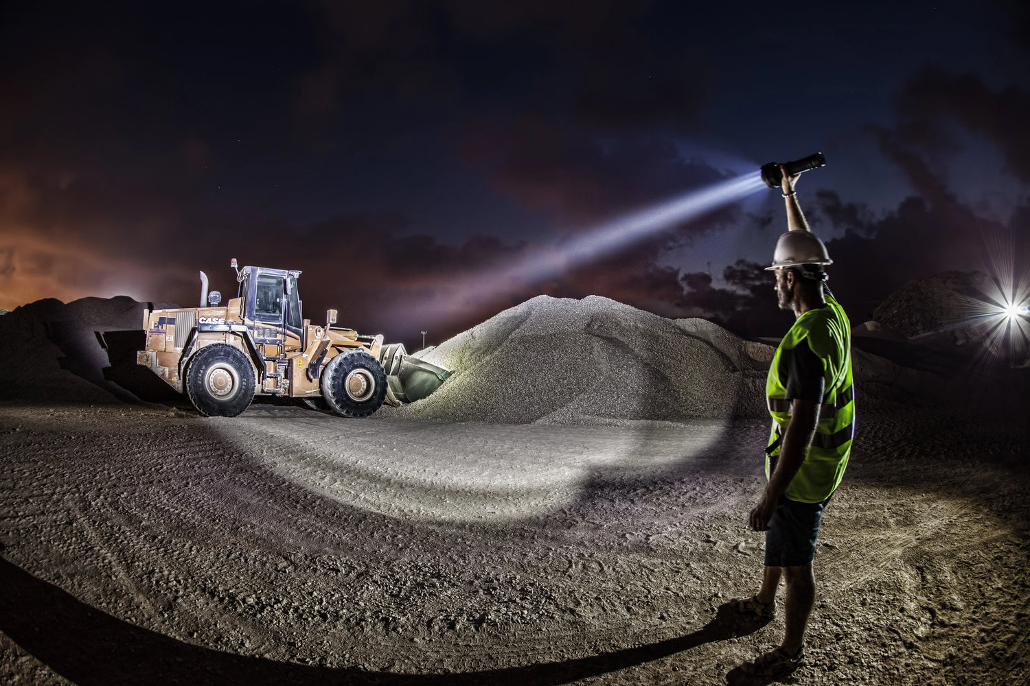 Baustellenarbeiter mit Helm, Warnweste und Stirnlampe repariert Baumaschine bei Nacht