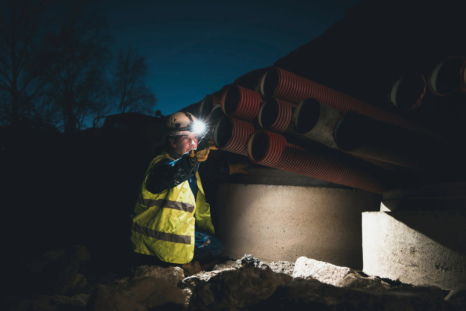 Baustellenarbeiter mit Helm, Warnweste und Stirnlampe repariert Baumaschine bei Nacht